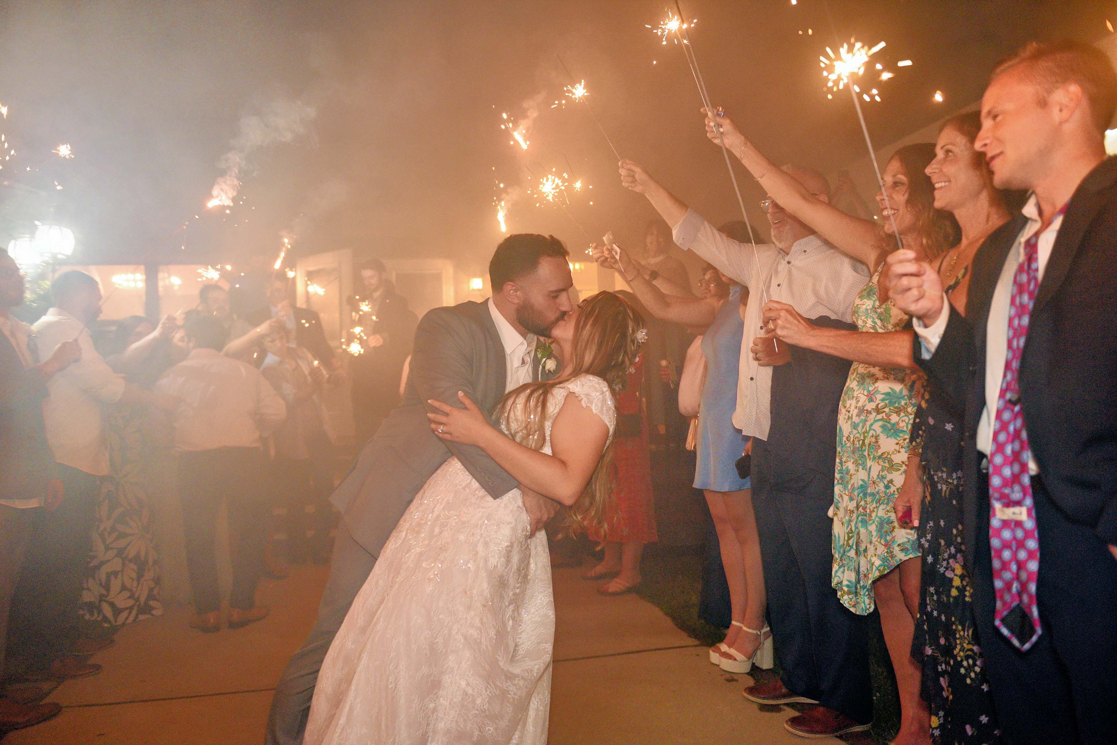 A newlywed couple kisses as friends celebrate with sparklers